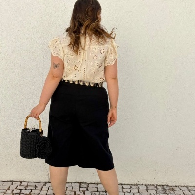 Pessoa de costas com camisa bege rendada, saia preta, e mala preta de cesto com alça de bambu