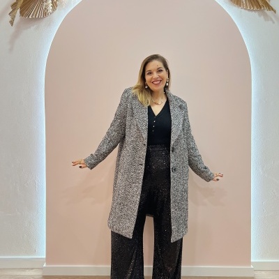 Mulher com casaco comprido cinza espinha de peixe e calças pretas brilhantes em fundo rosa com arco e decoração seca.