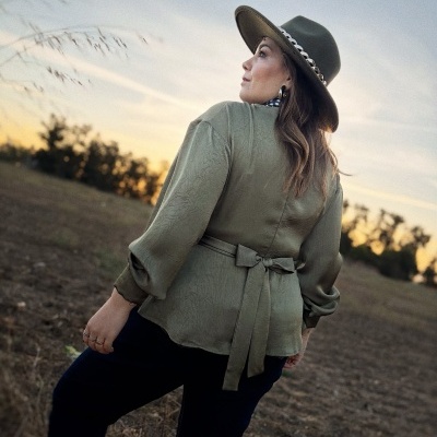 Mulher de costas vestindo blusa verde com laço e chapéu verde com corrente prata ao pôr do sol.