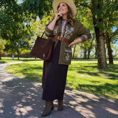 Mulher com casaco bordado, vestido castanho e chapéu bege num parque verde