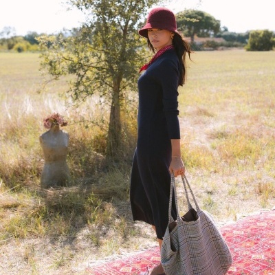 Mulher com vestido azul, chapéu bordô e bolsa xadrez, em campo aberto com tapete vermelho
