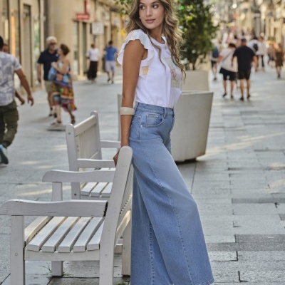 Mulher com t-shirt branca, calças de ganga largas azuis e sapatos dourados encostada a banco branco numa rua pedonal