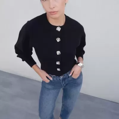 Mulher com camisola preta abotoada, jeans azuis e sapatos mule xadrez em fundo cinza