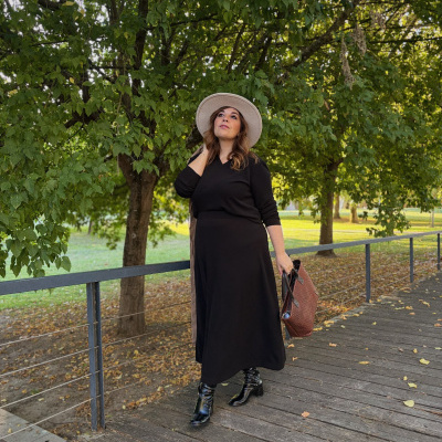 Mulher com roupa preta, botas e chapéu bege num passadiço de madeira ao ar livre perto de árvores.