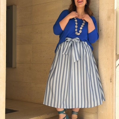 Mulher com camisola azul, saia às riscas azuis e brancas e sandálias verdes em escadas de pedra