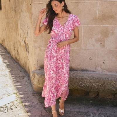 Mulher com vestido rosa e branco comprido, sandálias douradas, ao ar livre junto a parede de pedra.