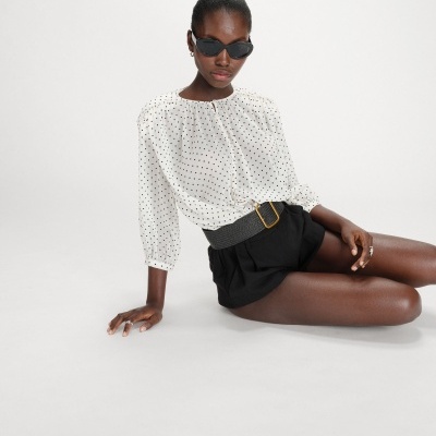 Mulher com blusa branca de polka dots e calções pretos com cinto cinzento, sentada em fundo branco