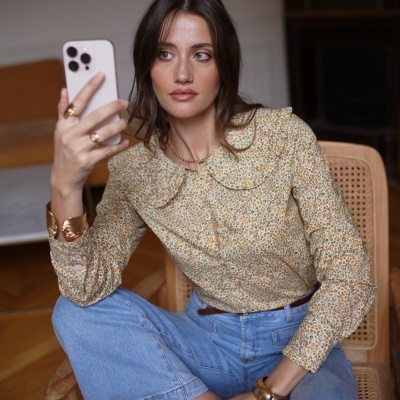 Mulher vestindo blusa floral e calças de ganga sentada numa cadeira de madeira, a usar acessórios dourados e a olhar para um smartphone branco