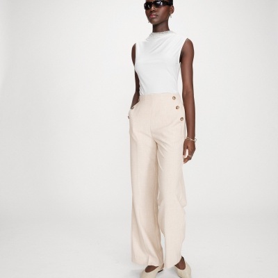 Mulher vestindo blusa branca, calças largas bege com botões, sapatos bege e óculos escuros.