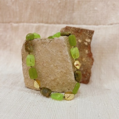 Pulseira verde e dourada com contas geométricas colocada em pedras sobre tecido bege