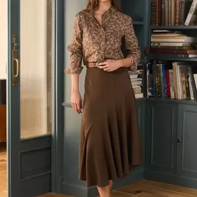 Mulher com camisa floral e saia castanha em ambiente interior com estante e porta azul