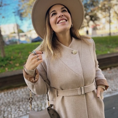 Mulher com casaco bege claro, chapéu bege e mala bege na zona urbana exterior.