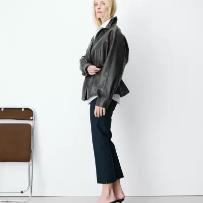 Mulher com casaco preto de couro, camisa branca, calças pretas e sandálias de salto em fundo branco.