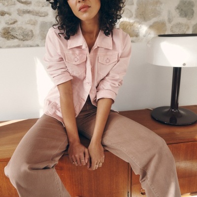 Mulher sentada usando casaco rosa claro de tecido denim com botões, calças largas bege, perto de parede de pedra e lâmpada.