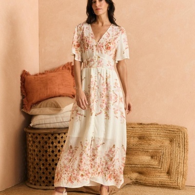 Mulher vestindo vestido longo floral branco com sapatos de cunha rosa, posando em ambiente com almofadas e tapete de fibras naturais.