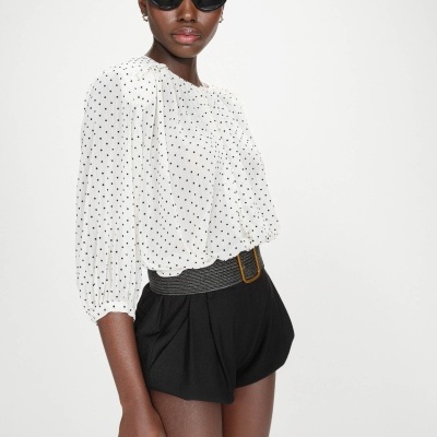 Modelo a usar blusa branca com bolinhas pretas, calções pretos com cinto e óculos de sol pretos em fundo branco