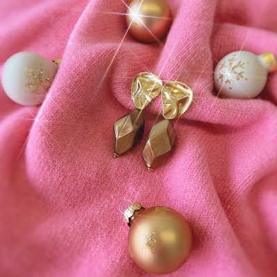 Brincos dourados geométricos sobre tecido rosa com bolas decorativas natalícias.