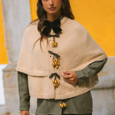 Mulher com capa bege de malha com ornamentos dourados e laço preto, camisa e calças verdes em ambiente exterior amarelo.