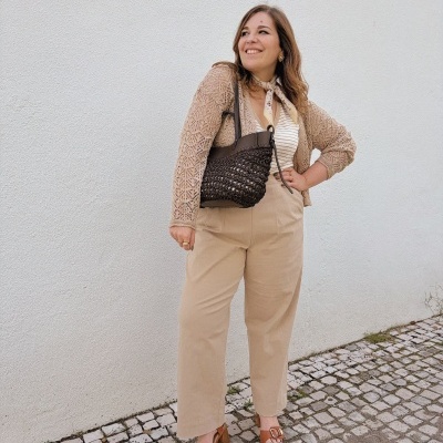 Mulher com roupa bege e mala castanha em piso de calçada com parede branca ao fundo