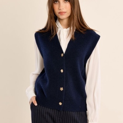 Mulher com colete azul escuro com botões dourados e camisa branca com calças risca de giz pretas.