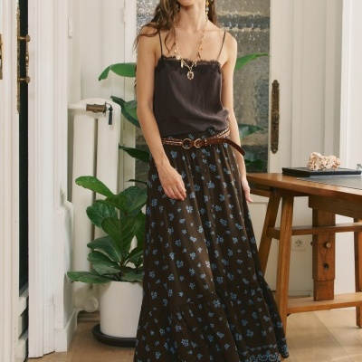 Mulher a usar vestido longo castanho com flores azuis e top preto de alças finas em interior com planta e mesa de madeira