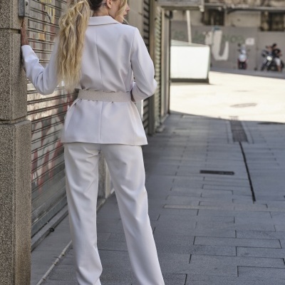 Mulher de costas veste fato branco com blazer e calças, sandálias bege, encostada num pilar junto a porta metálica