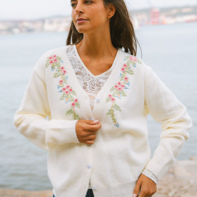 Casaco branco bordado com motivos florais coloridos e blusa branca com renda ao fundo marítimo.