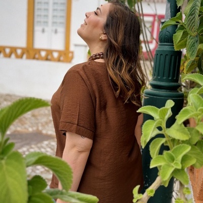 Mulher com blusa castanha de manga curta e colar castanho em cenário exterior com plantas e edifício