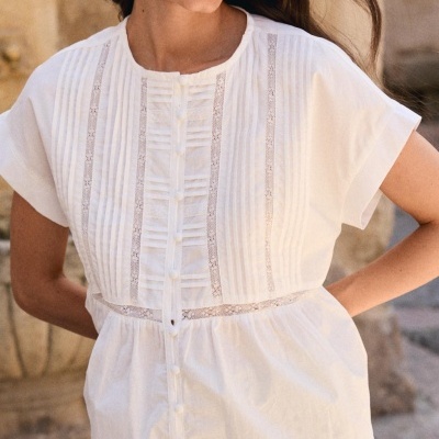 Blusa branca com pregas e renda, detalhes de botões, em mulher sorridente.