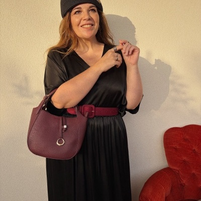 Mulher com vestido preto e cinto borgonha segura mala borgonha em pele, chapéu preto com detalhe dourado, poltrona vermelha ao lado.