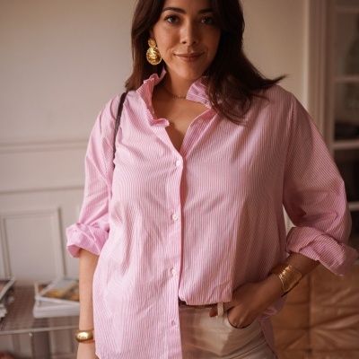 Mulher com camisa rosa às riscas brancas, calças claras e acessórios dourados num ambiente interior