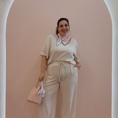 Mulher vestindo conjunto bege de linho com lenço rosa e mala rosa em fundo rosa com decoração floral seca