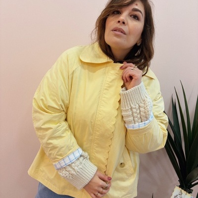 Mulher com casaco amarelo claro com detalhes ondulados e punhos de malha creme, jeans azuis, em frente a parede clara e planta verde.