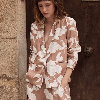 Conjunto feminino castanho claro com padrão floral branco, blazer e calças largas, modelo encostada à parede de pedra