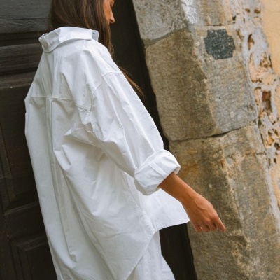Camisa branca de tecido leve com mangas dobradas ao lado de parede de pedra