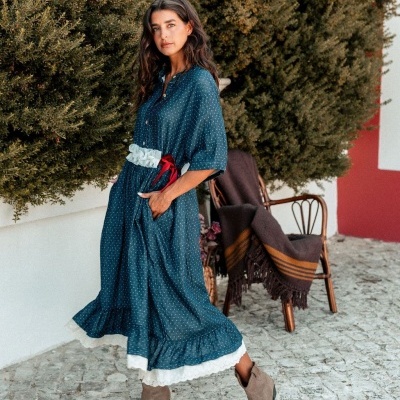 Vestido azul comprido com bolinhas brancas e cinto branco com laço vermelho, botas castanhas, cadeira e cobertor ao fundo