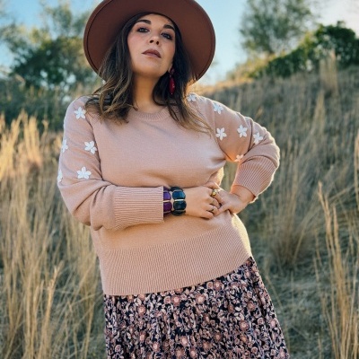 Mulher a usar camisola bege de flores brancas, saia floral azul e rosa e chapéu castanho em ambiente exterior com ervas secas
