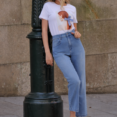 Mulher com t-shirt branca estampada, jeans azul e sapatos pretos encostada a poste verde na rua