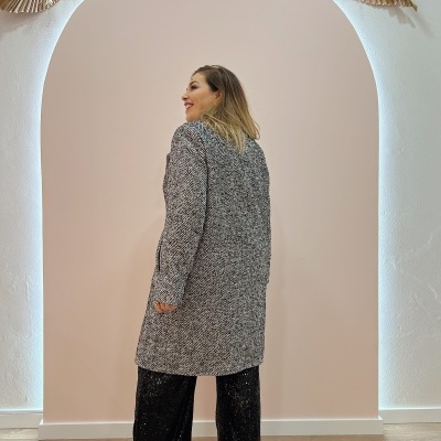 Mulher de perfil veste casaco cinza com padrão espinha de peixe em fundo interior claro com arco decorado.