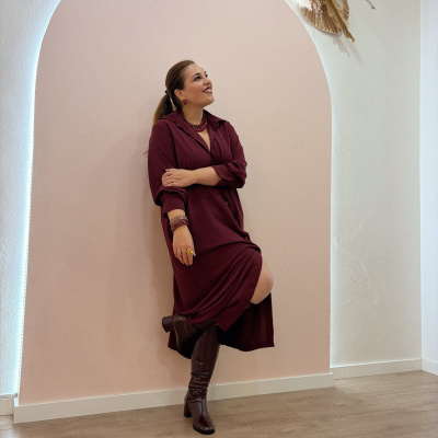 Mulher com vestido bordeaux, botas castanhas, parede rosa e branca com decoração em folhas secas