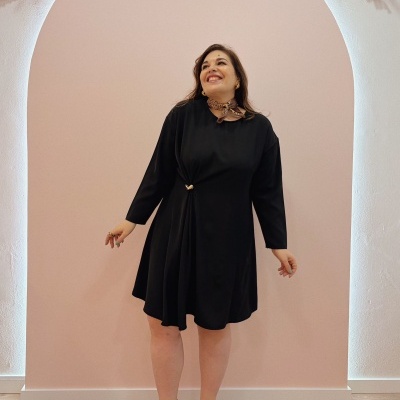 Mulher de vestido preto com detalhe na cintura, sandálias marrons e lenço ao pescoço, em frente a parede rosa com decoração seca