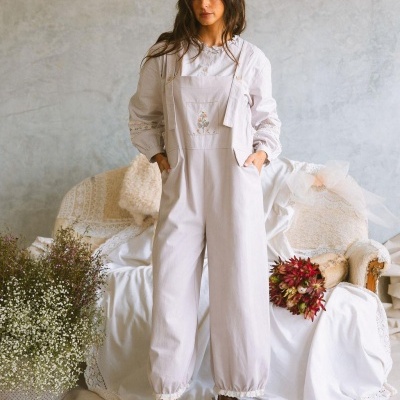 Mulher com macacão bege claro com detalhe bordado, em ambiente com sofá e flores