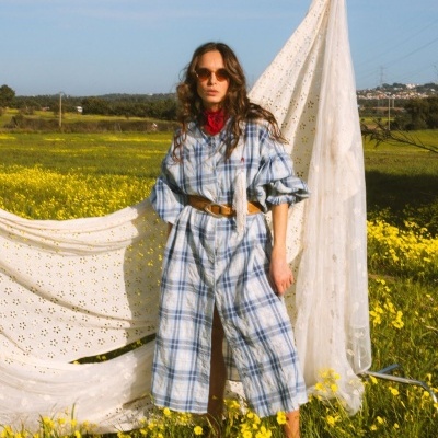 Mulher com vestido xadrez azul e branco, cinto castanho, lenço vermelho, óculos escuros em campo com flores amarelas e tecido rendado.
