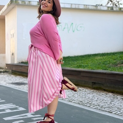 Mulher sorridente de chapéu e roupa rosa numa rua urbana
