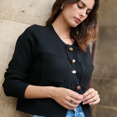 Casaco feminino preto com botões dourados e bolsos, usado com calças de ganga azuis claras próximo a parede de pedra
