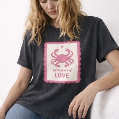 Mulher a usar t-shirt preta com estampado de caranguejo rosa e texto em inglês sentado num sofá branco