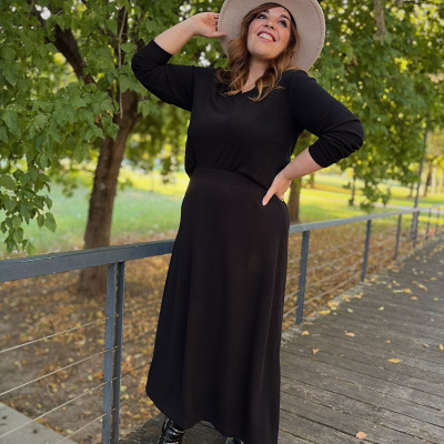 Mulher a usar vestido preto comprido, botas pretas e chapéu bege num passadiço de madeira com árvores ao fundo