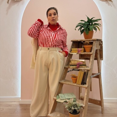 Pessoa veste camisa riscada vermelha e branca, calças bege e botas castanhas ao lado de escada com plantas e livros.