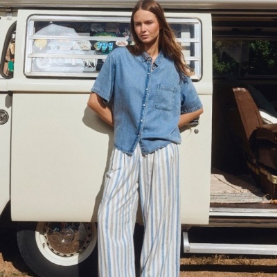 Mulher com camisa de ganga azul e calças largas ás riscas junto a carrinha antiga branca