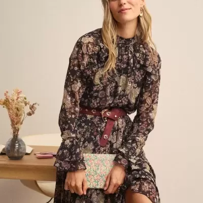 Mulher com vestido floral e cinto bordô sentada segurando carteira floral perto de mesa com flores secas